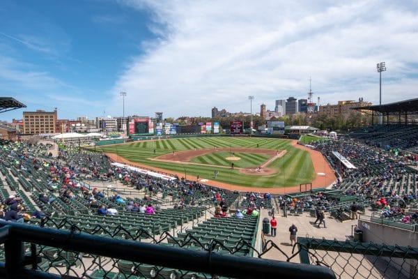 Wide view of a baseball stadium with green grass, infield dirt, and partially filled stands under a blue sky. City buildings are visible in the background beyond the outfield—an ideal stop for any Rochester Overnight Itinerary.