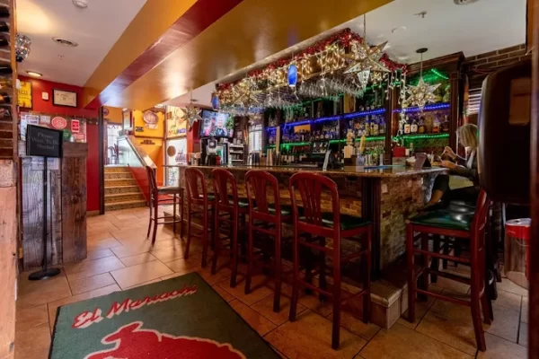 A cozy bar with wooden stools, colorful lights, and festive decorations—just the place to unwind after an overnight in Albany. Bottles line the shelves behind the bar as a person sits at the counter, while a red mat with text greets guests near the entrance. The tiled floor completes this inviting atmosphere.