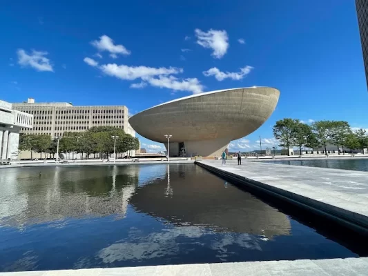 A futuristic, bowl-shaped building stands by a reflective pool under a blue sky with scattered clouds in Albany, surrounded by modern architecture and trees—the perfect backdrop for an unforgettable overnight in Albany.