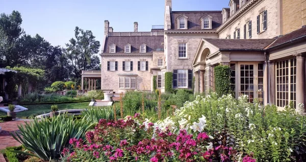 A large, elegant mansion with beige brick walls is surrounded by lush gardens filled with colorful flowers, green shrubs, and a brick walkway on a sunny day—an idyllic stop for your Rochester Overnight Itinerary.
