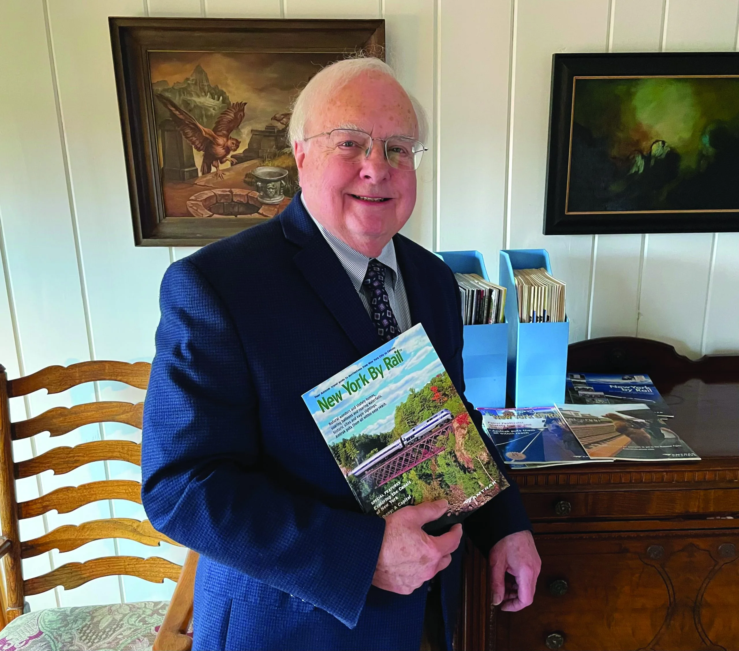 An older man in a suit and tie smiles while holding a “New York By Rail” magazine—perhaps reminiscing about The One That Got Away. He stands indoors beside a wooden table with magazines and file folders, while framed paintings hang on the white wall behind him.