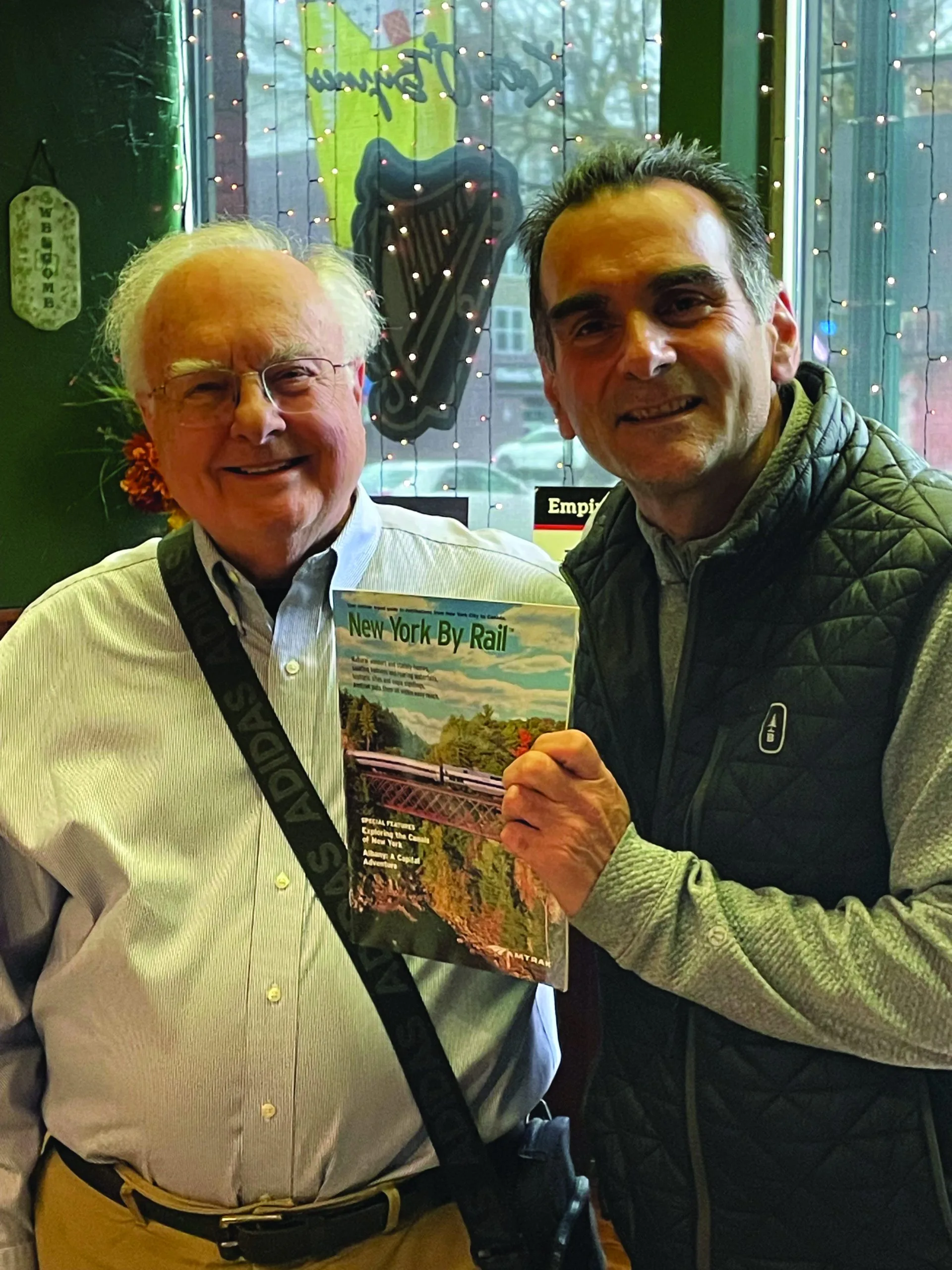 Two smiling men stand indoors. One holds a New York By Rail magazine. They are in front of a window decorated with string lights and Irish-themed decor, sharing a moment reminiscent of The One That Got Away.