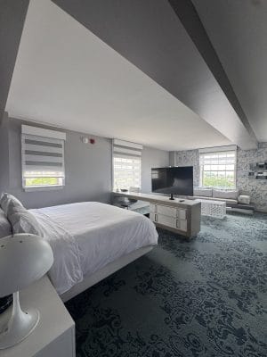 A modern hotel room with a large bed, white bedding, a flat-screen TV on a stand, patterned carpet, and two windows with striped blinds offers minimalist decor in shades of gray and white—the perfect retreat after an adventurous day exploring Hudson River Cruises.