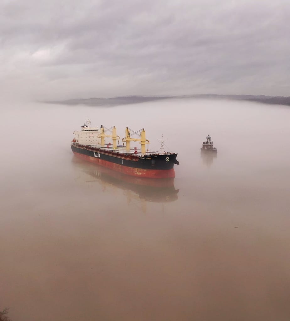 A large cargo ship and a small lighthouse appear to float in dense fog over calm water, blending with the muted sky, evoking the ethereal ambiance of an Osheaga morning.