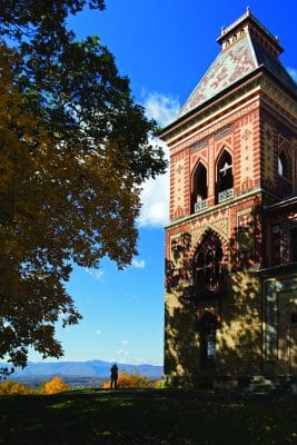 A tall, ornate brick tower stands beside autumn trees, with a person in the distance and mountains under a bright blue sky in the background—capturing the tranquil charm of an overnight in Hudson.