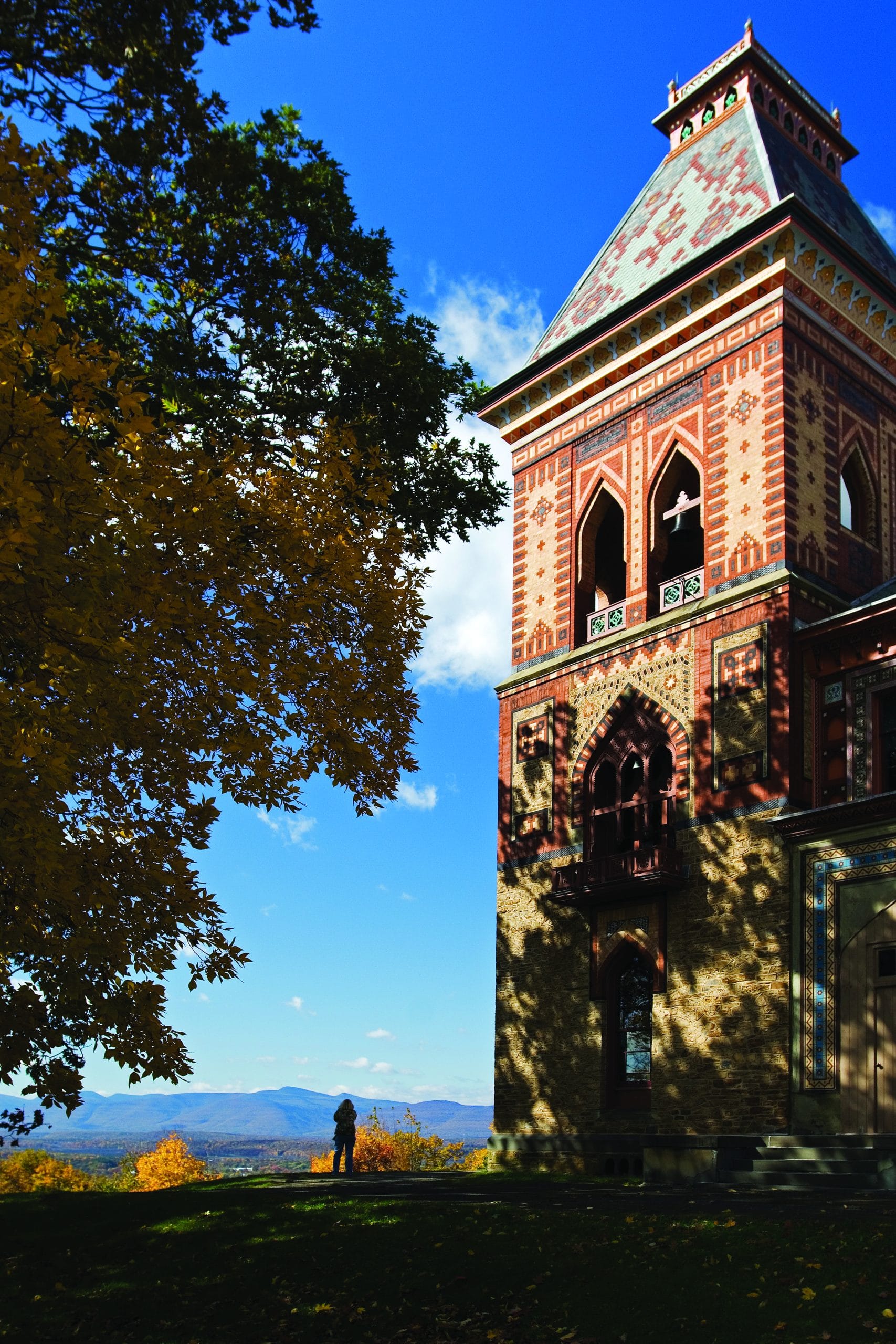 A tall, ornate brick tower stands beside autumn trees, with a person in the distance and mountains under a bright blue sky in the background—capturing the tranquil charm of an overnight in Hudson.