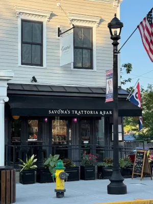 Front view of Savonas Trattoria & Bar with black awning, potted plants, an outdoor patio, a streetlamp, and an American flag. Perfect for visitors enjoying an overnight in Hudson, the scene is completed by a yellow fire hydrant on the sidewalk in front.
