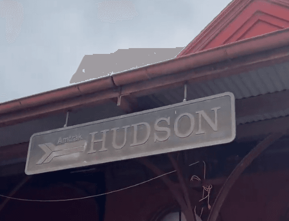 A weathered sign reading Amtrak HUDSON hangs under a metal roof at a train station, hinting at the possibility of an overnight in Hudson, with part of a red building and cloudy sky visible in the background.