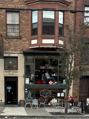 A street view of a café called Daily Grind with outdoor seating, neon signs, and a person sitting in the large front window above the entrance—perhaps unwinding after spending the night Overnight in Albany. A tree partially obscures the front.
