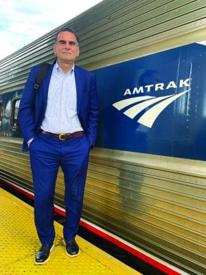 A man in a blue suit stands on a yellow platform next to an Amtrak train, with the Amtrak logo visible on the train car behind him—an unmistakable scene for any traveler riding New York by Rail.