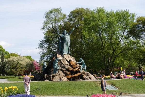 Bronze statue of a robed figure with raised arms atop rocks in a park, surrounded by smaller statues and blooming flowers, with people walking and relaxing nearby under trees—an inviting scene for those enjoying an overnight in Albany.
