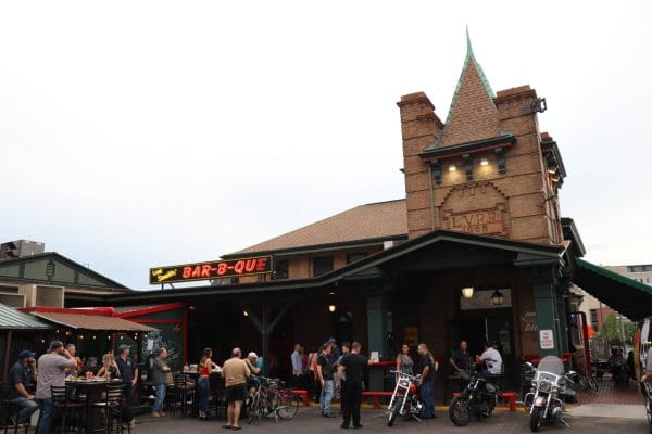 People gather outside a historic brick building with motorcycles parked nearby; a neon BAR-B-QUE sign hangs over the entrance, and diners sit at outdoor tables under a covered patio—the perfect spot to savor a meal during your Rochester Overnight Itinerary.