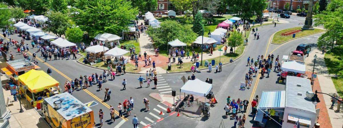 Aerial view of the vibrant LARAC June Arts Festival, showcasing a busy outdoor market with white tents, food trucks, and crowds walking along streets surrounded by green trees and brick buildings on a sunny day.
