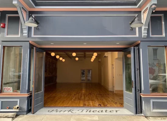 Open double doors reveal an empty theater space with wooden floors and round hanging lights; Park Theater is written on the entryway step, inviting you to imagine spending an overnight in Hudson surrounded by the charm of this unique venue.