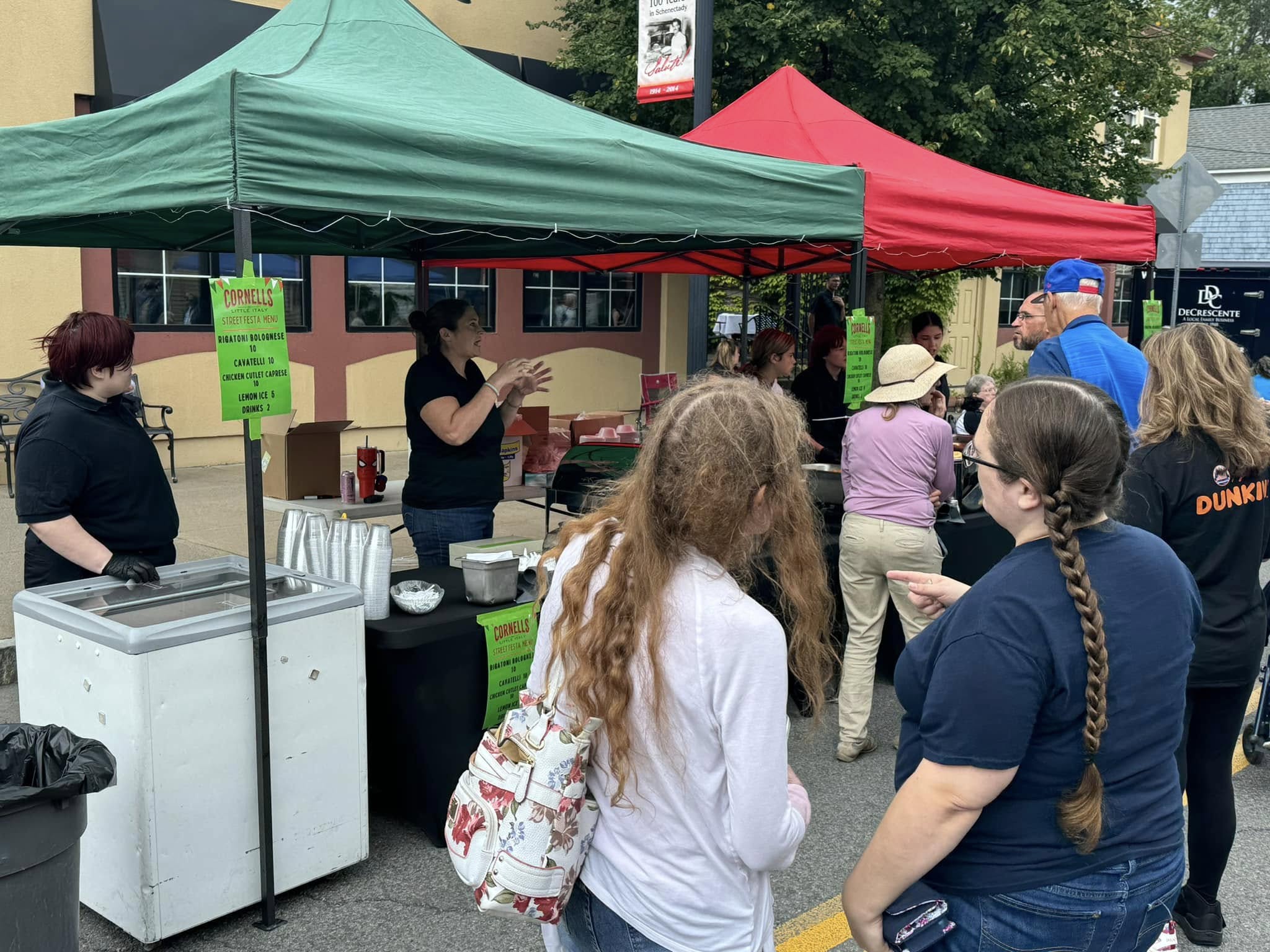 Several people stand in line at an outdoor market with green and red tents, engaging with vendors. A woman gestures while talking to the crowd, creating a lively atmosphere reminiscent of a Wine and Food Festival. Signs and coolers are visible on the tables under the tents.