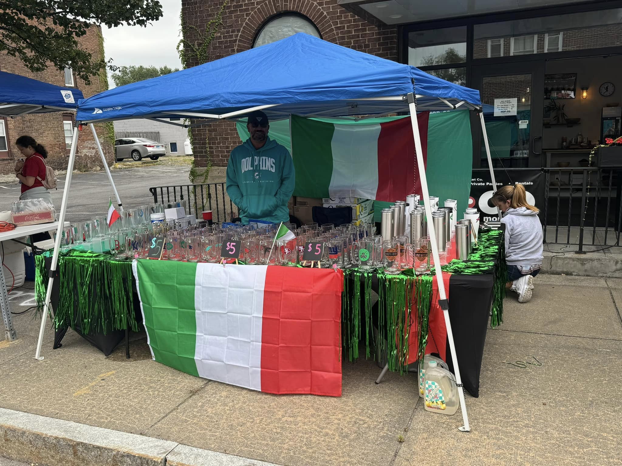 A vendor stands under a blue canopy with Italian flags, selling clear cups for $5 each at an outdoor market during the Wine and Food Festival. Green, white, and red decorations cover the table, with a large Italian flag draped in front.