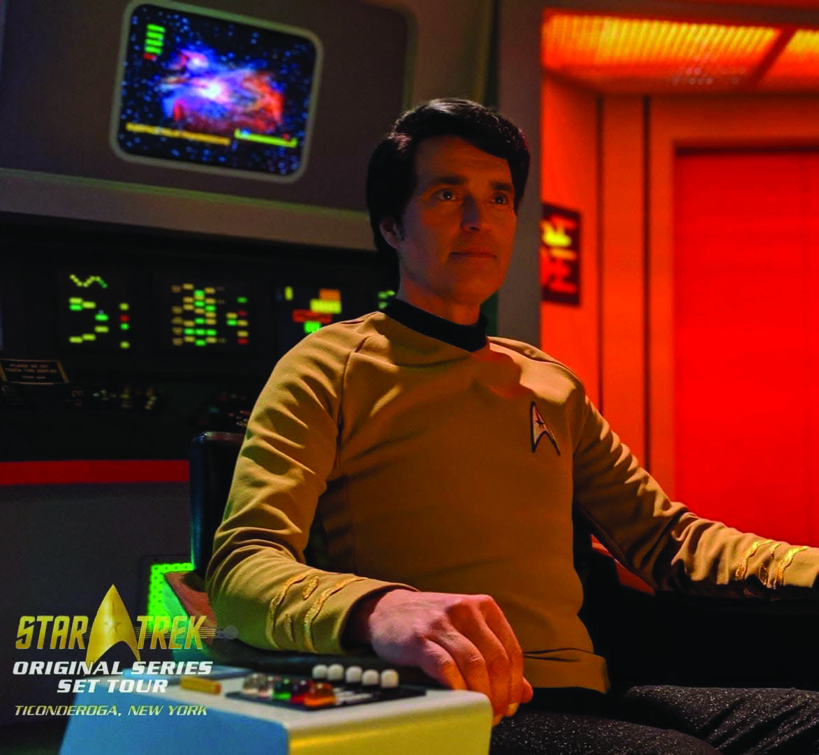 A person in a gold Star Trek uniform sits at the command chair of a starship bridge set, surrounded by futuristic controls and colorful screens. Text reads Star Trek Original Series Set Tour, Ticonderoga, New York, inviting fans to step onto the authentic Star Trek Original Series Set and experience the iconic bridge firsthand.