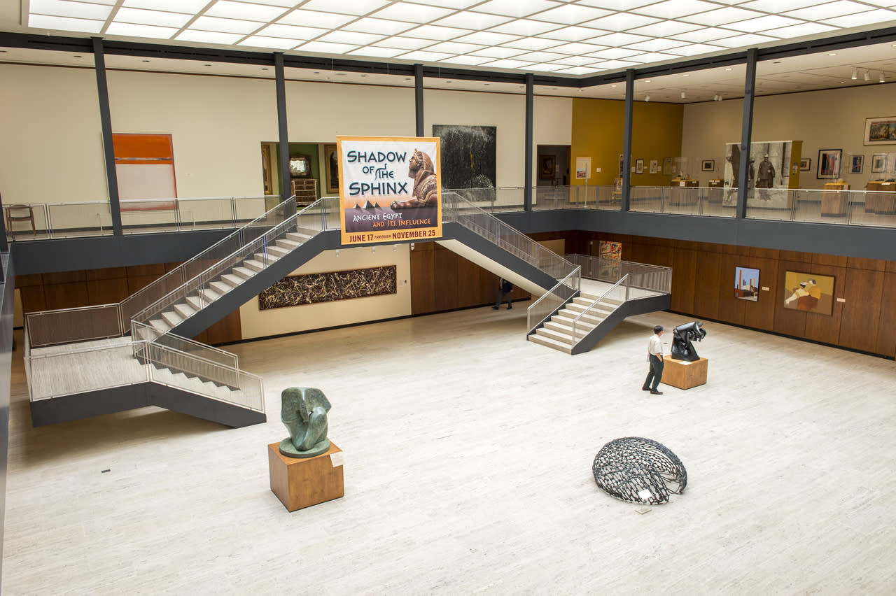 Spacious Munson art museum lobby with a central staircase, sculptures on the floor, paintings on the walls, and a large Shadow of the Sphinx exhibit banner above the stairs. Two people view the art.