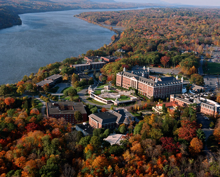 Aerial view of a college campus surrounded by colorful autumn trees, with a large river running alongside the campus buildings, evoking the sense of The One That Got Away as golden leaves drift gently past memories that linger on the water’s edge.