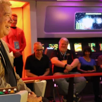 A smiling man sits on a control chair in a colorful Star Trek Original Series Set, while a group of people seated behind him watch and smile. A coffee cup is on the console in the foreground.