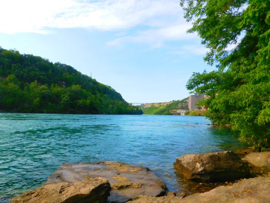 A river flows between green hills under a blue sky, with rocks in the foreground and trees lining the right side. In the distance on the right bank, near where the landscape opens up, sits a building reminiscent of those found at Rosamond Gifford Zoo.