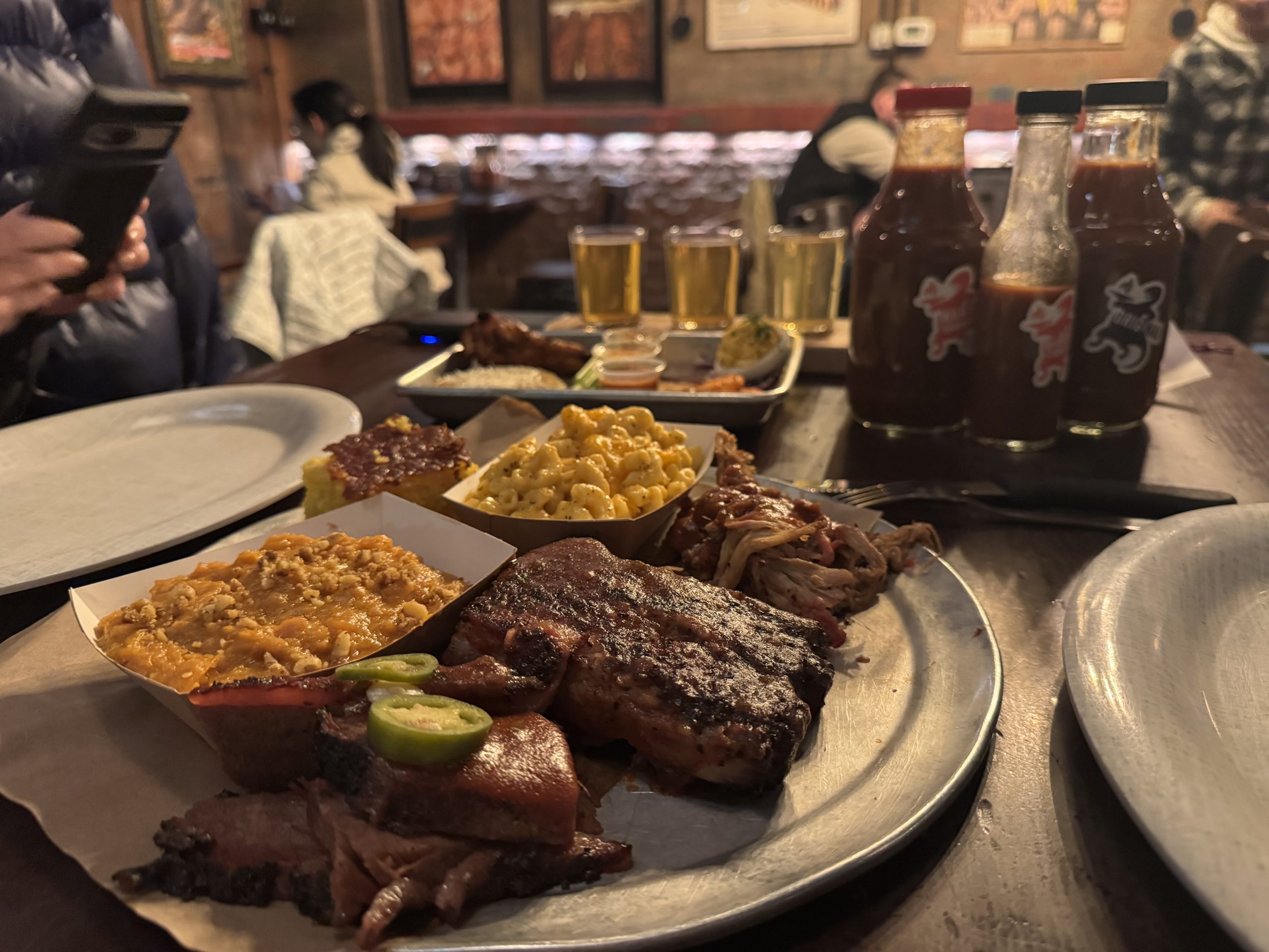 A metal plate with ribs, brisket, mac and cheese, baked beans, and cornbread sits on a wooden table at The One That Got Away. Behind are three bottles of BBQ sauce and a flight of beers. Diners sit in the warmly lit restaurant background.