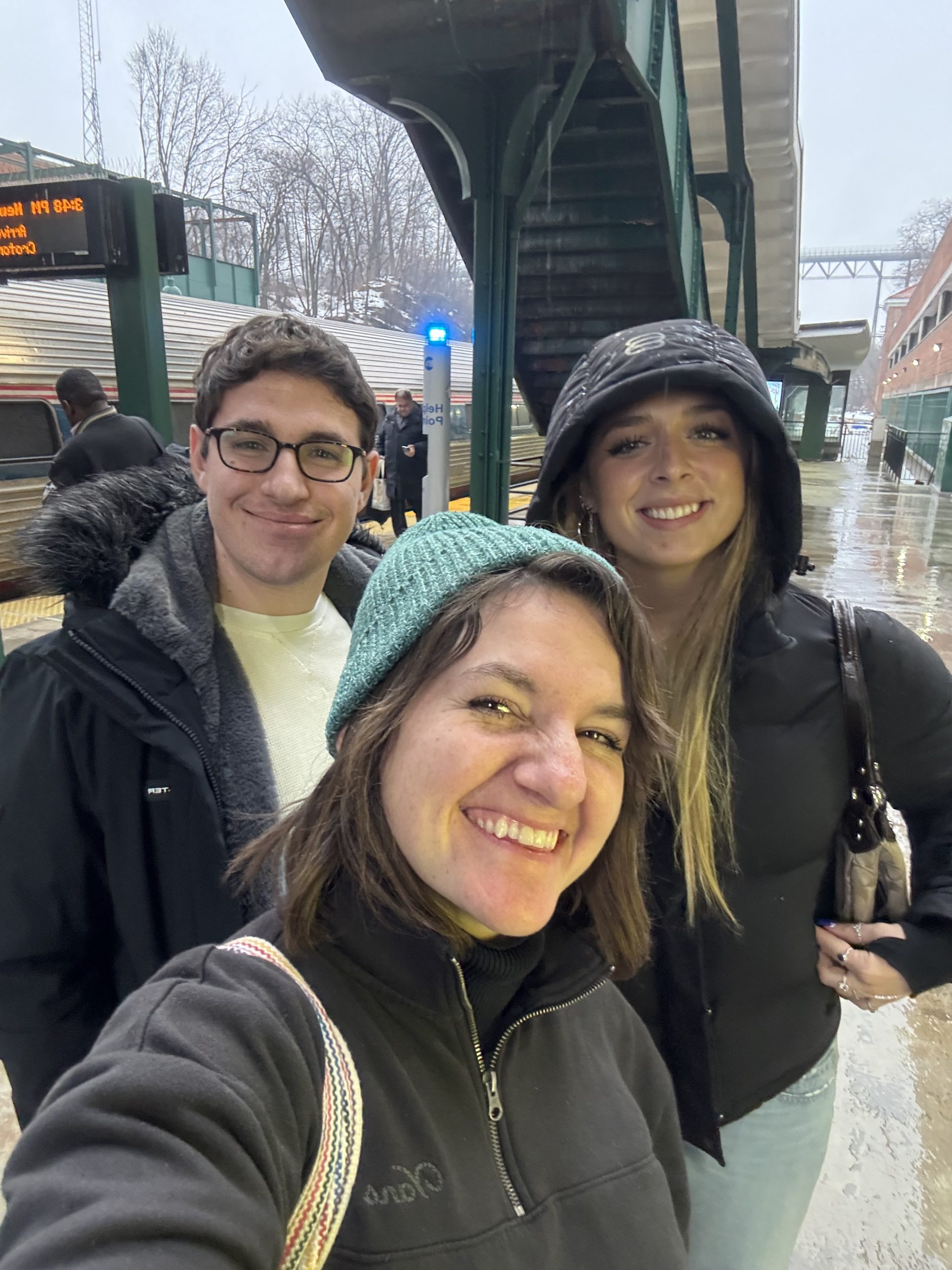 Three people stand and smile for a selfie at a train station on a rainy day, reminiscing about The One That Got Away. A train, wet platform, and digital sign are visible in the background as everyone stays warm in jackets and hats.