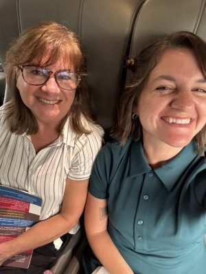 Two women smiling while sitting next to each other on a train or bus, sharing a warm moment that feels like it’s straight from The One That Got Away. The woman on the left holds a colorful pamphlet and wears glasses with a striped shirt, while the woman on the right, dressed in a teal shirt, laughs beside her.