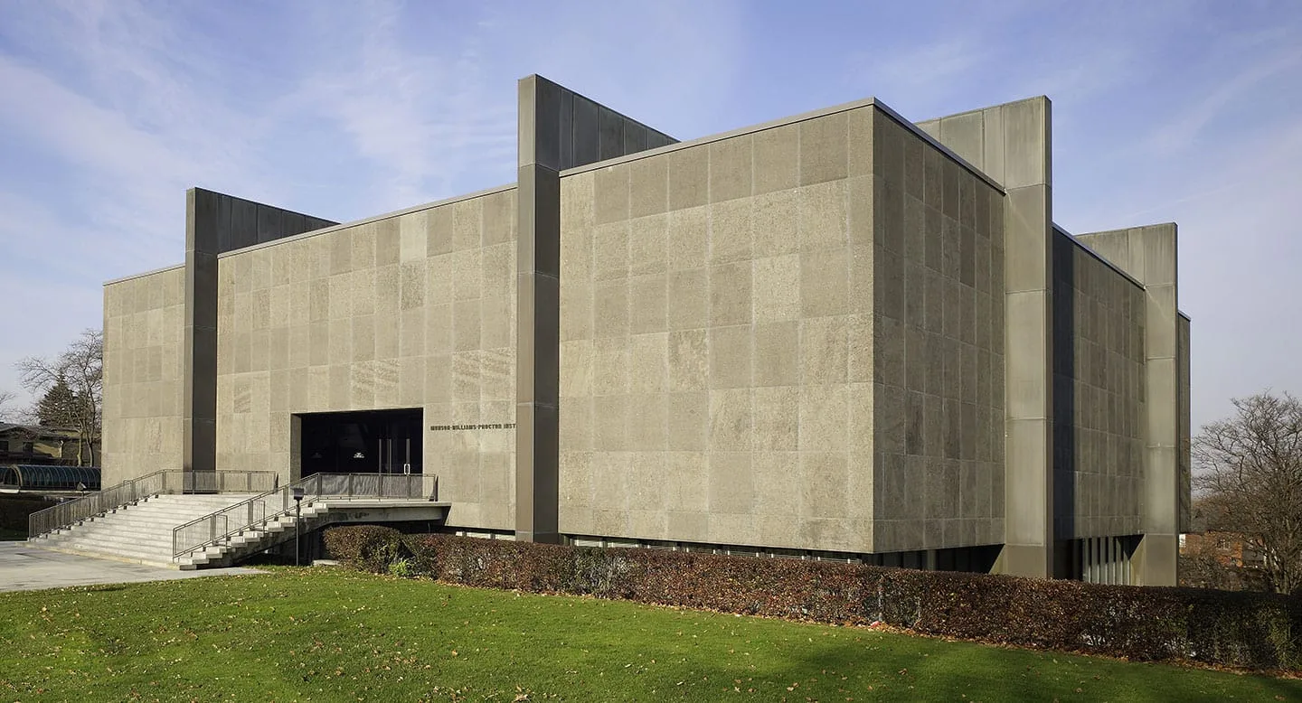 A large, rectangular modern building with tall, smooth concrete walls, small windows, and a staircase leading to a dark entrance stands surrounded by grass and a low hedge under a partly cloudy sky, reminiscent of the striking architecture found near Onondaga Lake State Park.