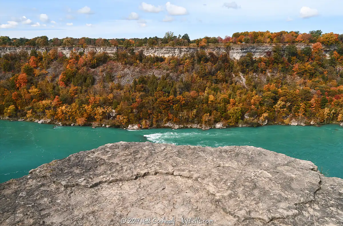 A rocky ledge in Devil’s Hole State Park overlooks turquoise water with white rapids. Behind the river, trees in autumn colors cover a steep, rocky cliff under a blue sky with scattered clouds.