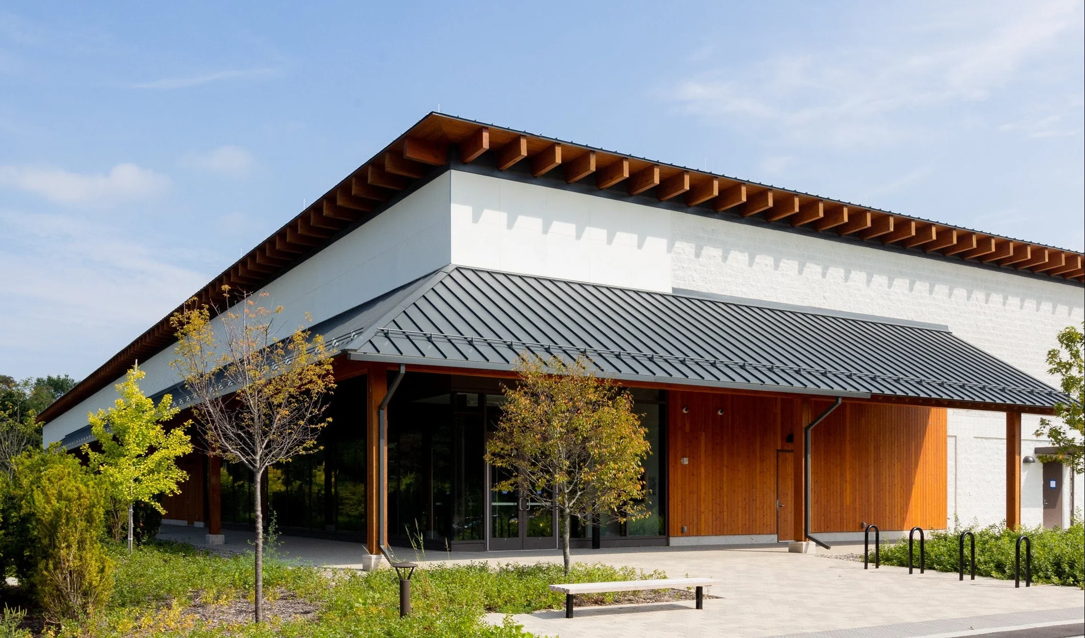 Modern building with wood paneling, large windows, and a metal roof surrounded by greenery and young trees under a sunny blue sky—reminiscent of the tranquil setting at Dia Beacon. A wooden bench is on a stone path near the entrance.
