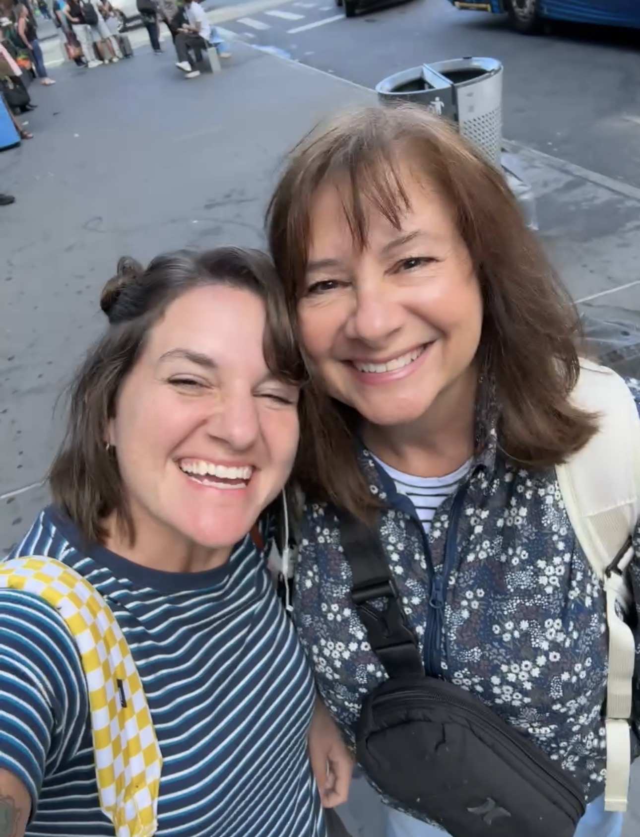 Two women, perhaps on a mother daughter Amtrak train trip, smile and pose together on a city sidewalk. One wears a blue striped shirt and yellow checkered backpack, the other a floral jacket with a black crossbody bag. People and vehicles are in the background.