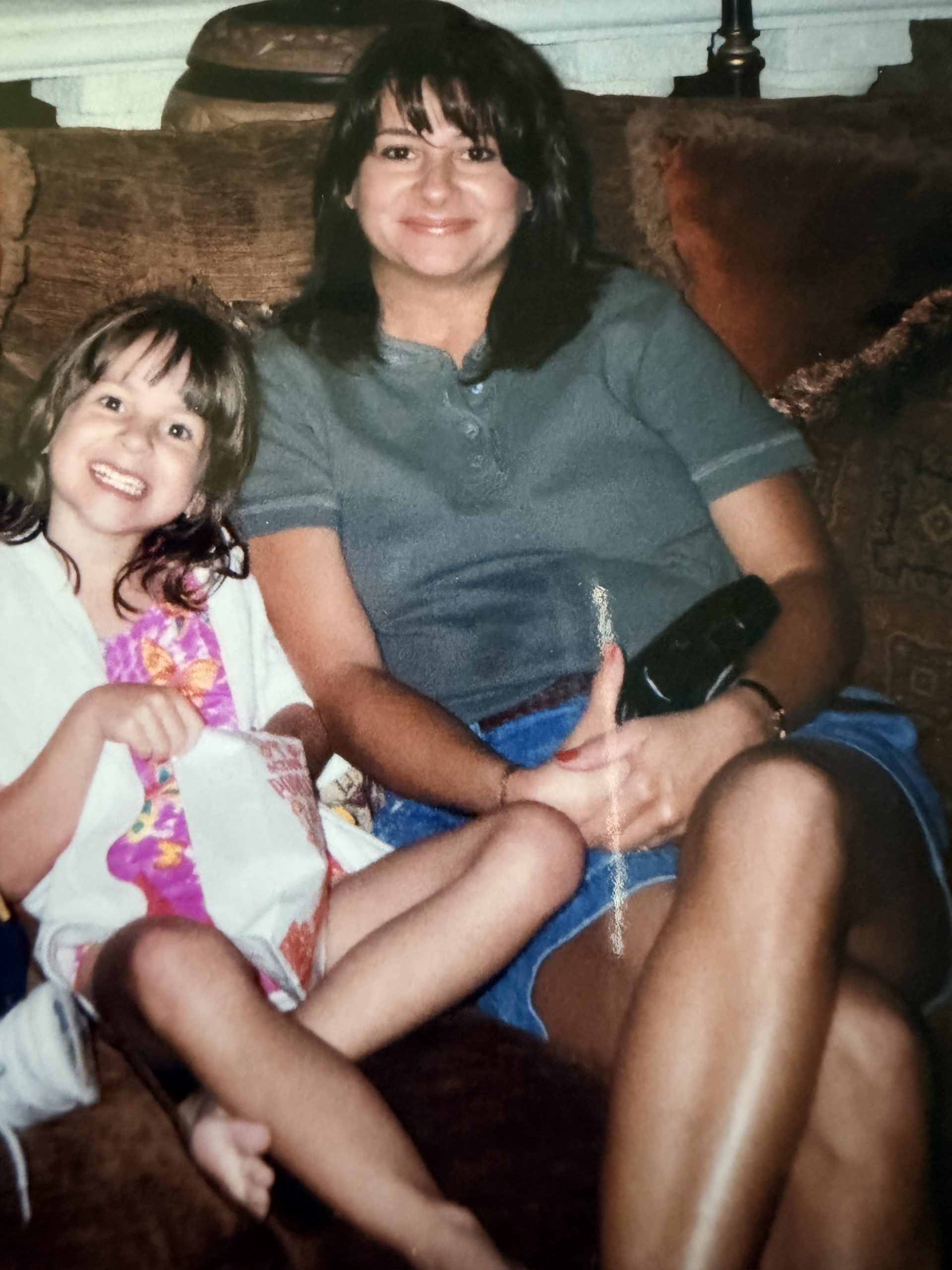 A mother and daughter sit closely together, smiling on a brown sofa. The girl holds a towel and wears a colorful dress, while her mother holds a remote and wears a gray shirt and blue shorts. Their joyful expressions capture the warmth of their bond, reminiscent of fond memories from their recent Amtrak train trip.