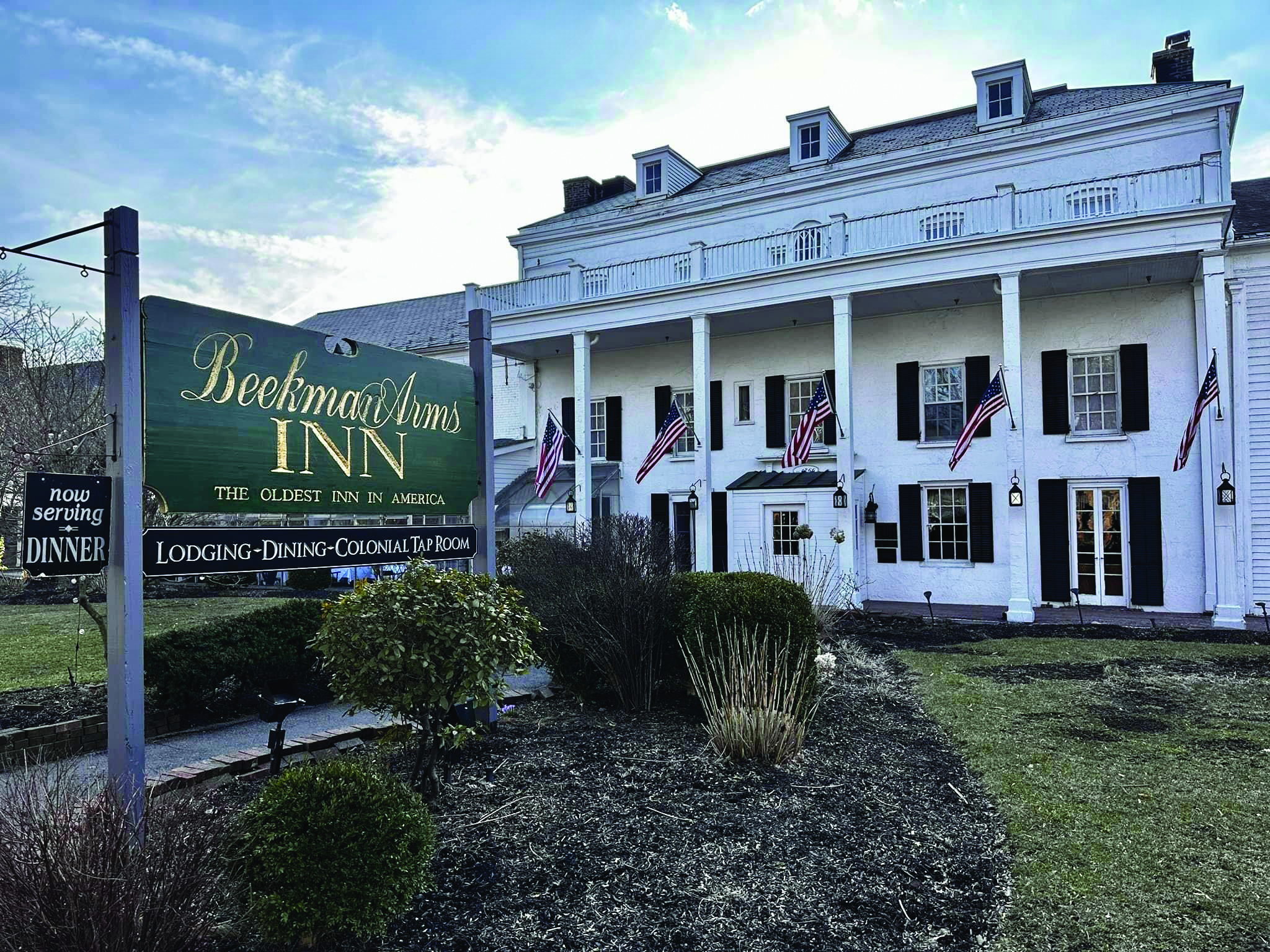 Historic white colonial inn with columns and American flags, set behind a green sign reading Beekman Arms Inn—The Oldest Inn in America. Neatly trimmed bushes and a lawn are in the foreground, evoking the charm of The One That Got Away—a timeless destination where history lingers and memories are made.