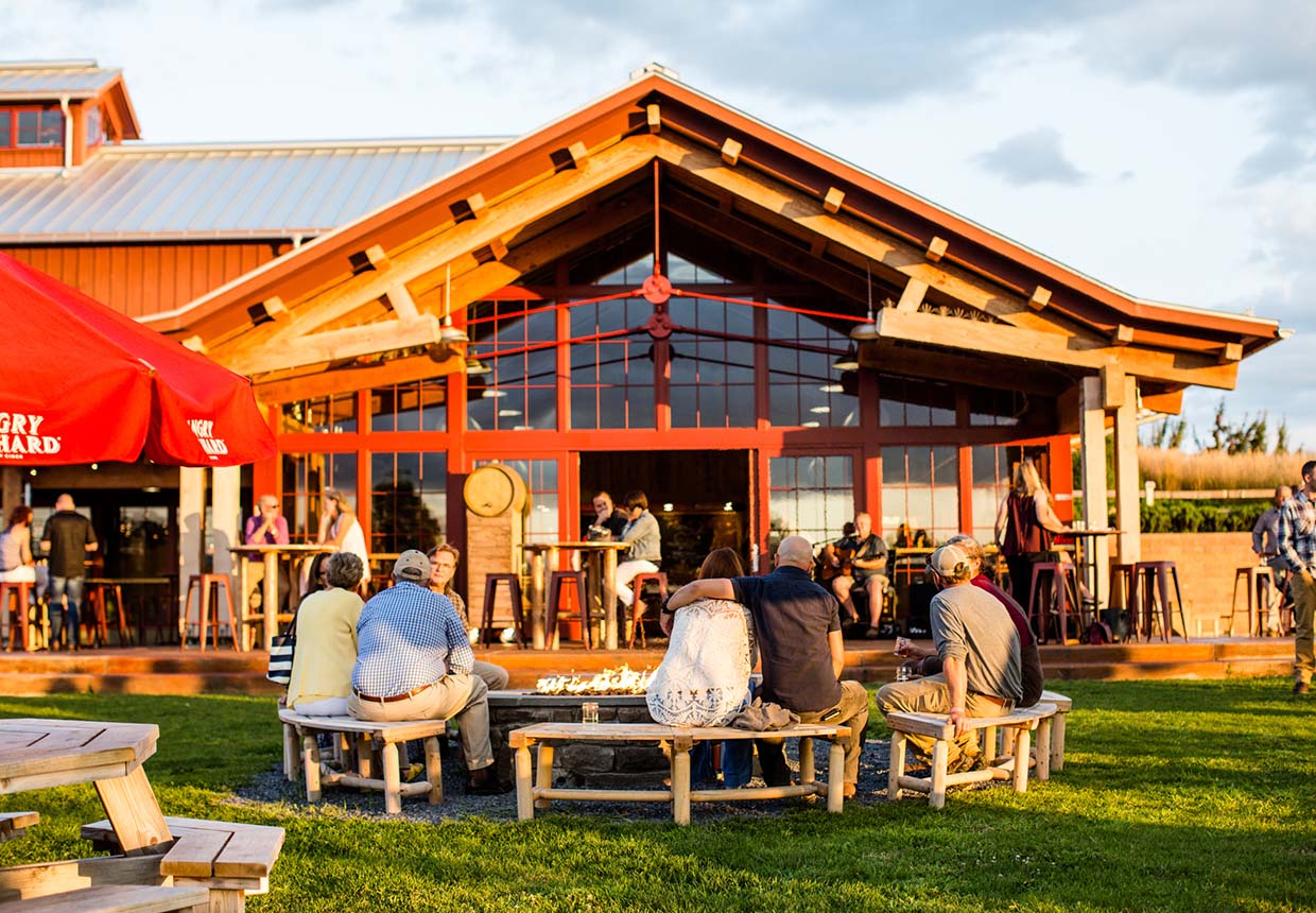 People sit around a fire pit outside the rustic wooden Cider House with large windows, enjoying a sunny day and sipping on refreshing hard cider from Angry Orchard. Others are seated at tables under a red umbrella and near the building. Grass and clear skies are visible.