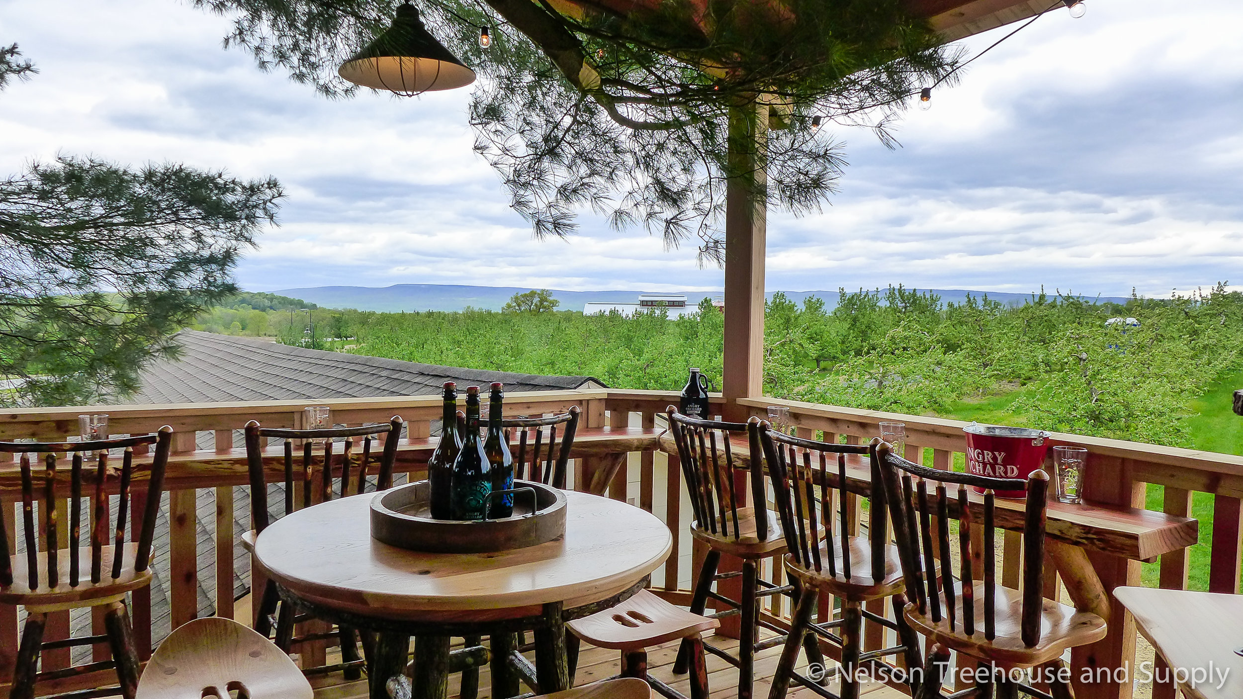 A wooden deck with chairs and a round table holding several bottles of hard cider overlooks a forested area under a cloudy sky. A hanging lamp and tree branches are visible above, evoking the relaxed ambiance of a Cider House like Angry Orchard.