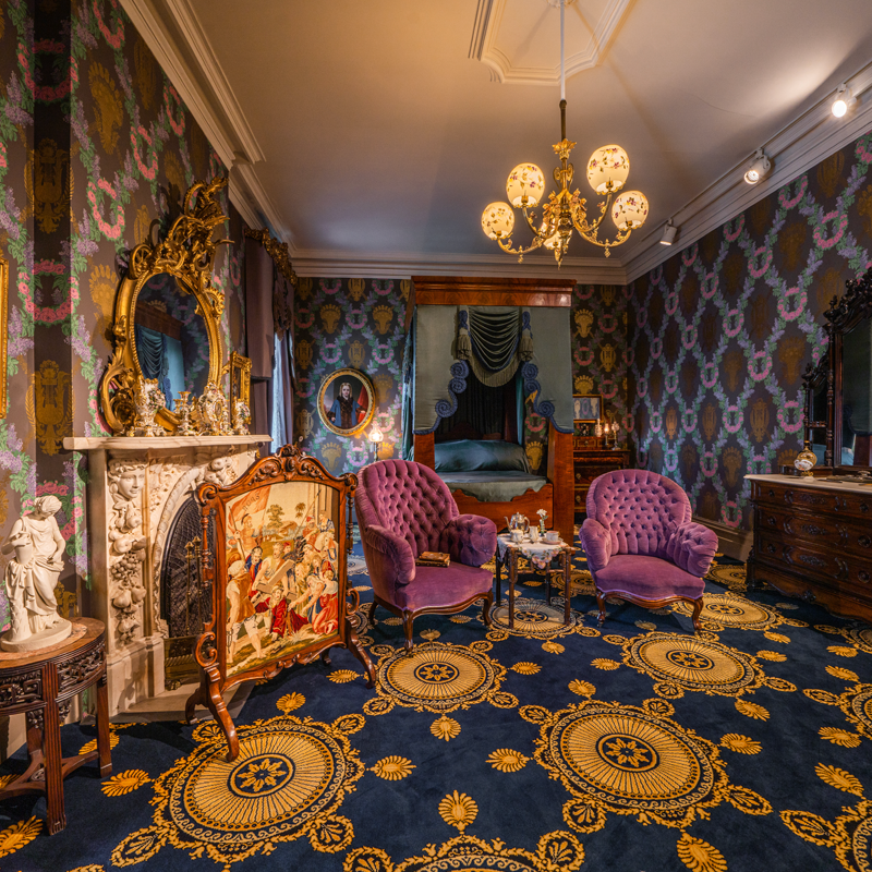 Ornate Victorian-style living room with two purple chairs, a marble fireplace, gold-framed mirror, patterned wallpaper, elaborate carpet, chandelier, and a view into a bedroom with a canopy bed—capturing the timeless elegance often found in Munson-inspired interiors.