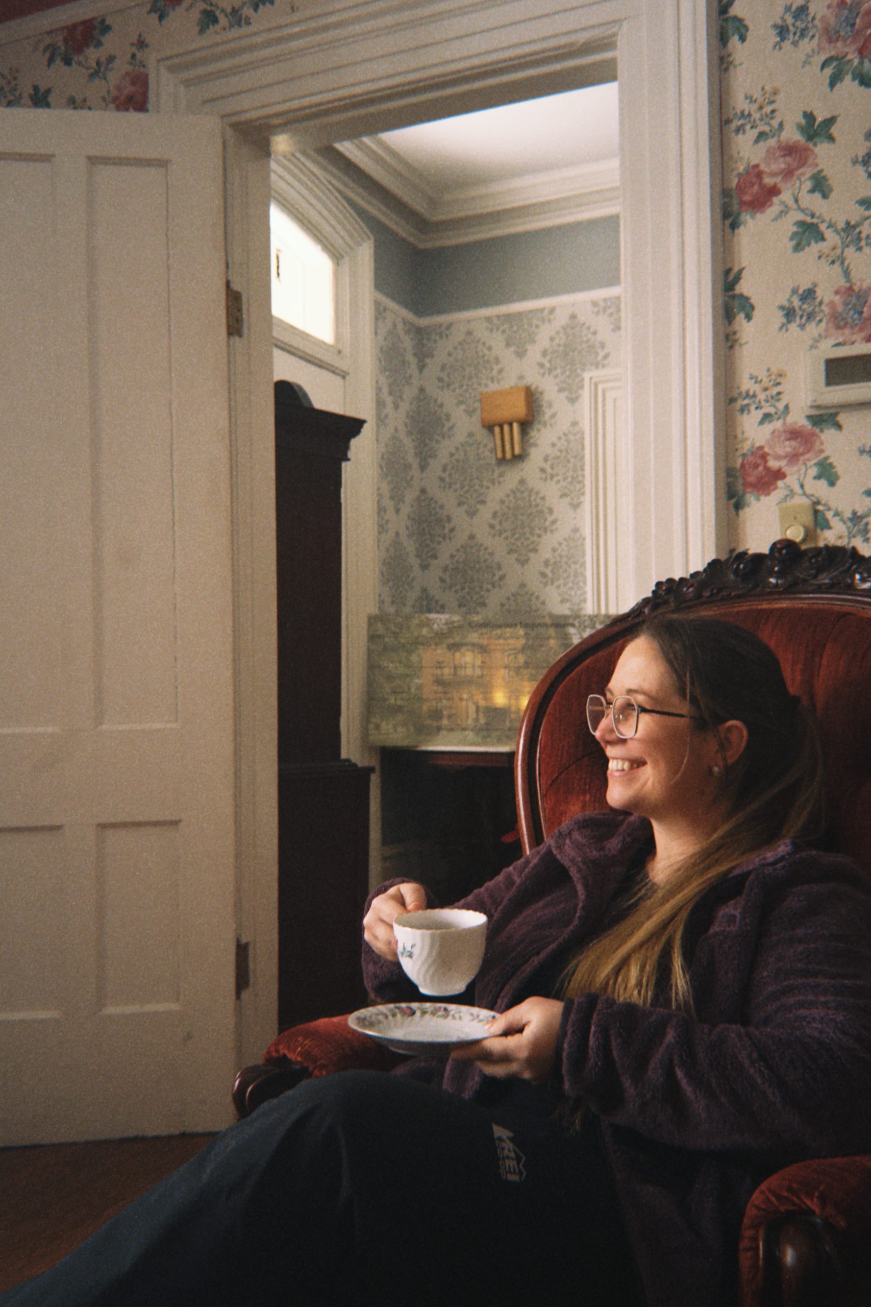 A woman with long hair and glasses sits smiling in a red armchair, holding a tea cup and saucer, in a vintage room with floral wallpaper and an open white door—her gaze carries the warmth of reminiscence, as if she’s pondering The One That Got Away.