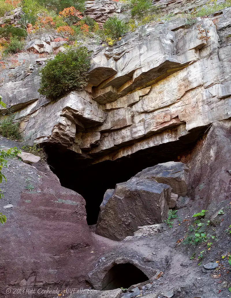 A rocky cave entrance with jagged stone overhangs and some greenery growing atop and around the rocks, reminiscent of the rugged beauty found in Devil’s Hole State Park. The cave opens into darkness below layered cliffs.