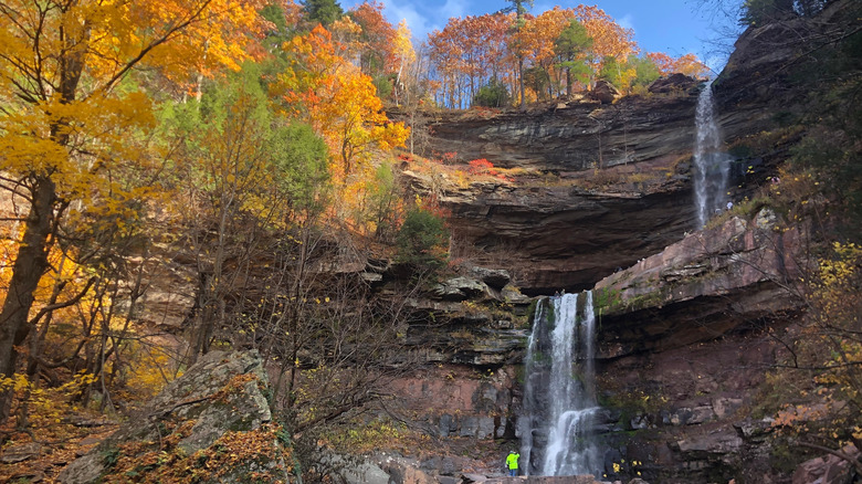 A person in a bright jacket stands near a two-tiered waterfall, part of the scenic Hudson River School Art Trail, surrounded by rocky cliffs and colorful autumn trees under a blue sky.