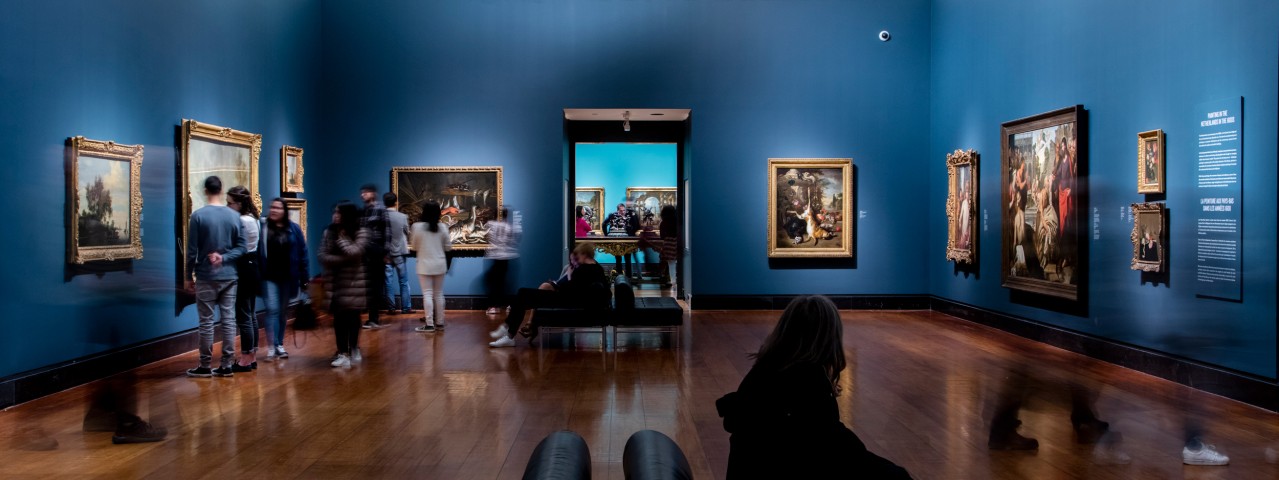 People view framed paintings on blue walls in a museum gallery, reminiscent of the tranquil atmosphere at the Art Gallery of Ontario. Some figures appear blurred, suggesting movement, while others sit or stand quietly observing the art. Polished wooden floors reflect the lighting.