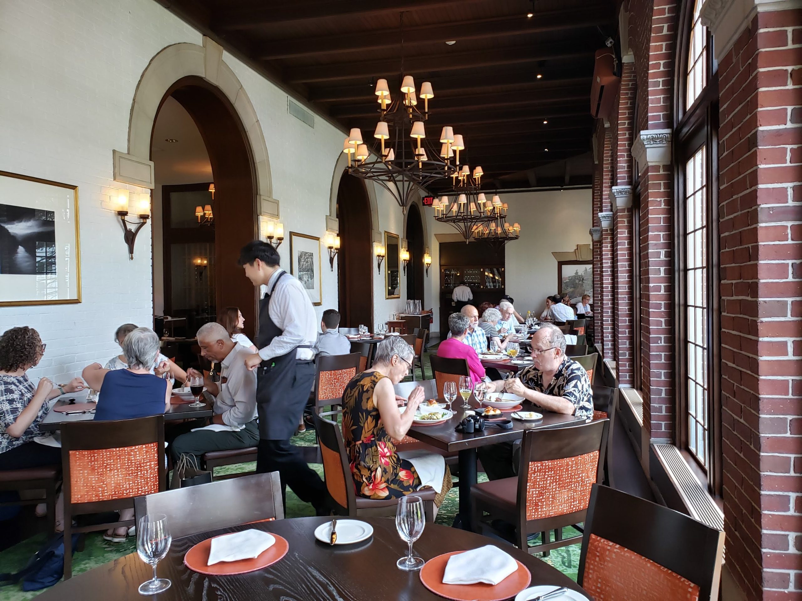 A busy, elegant restaurant—reminiscent of the renowned American Bounty Restaurant—with people dining at tables. Large arched windows line one wall, and chandeliers hang from a dark wooden ceiling. A waiter serves guests as natural light fills the room.