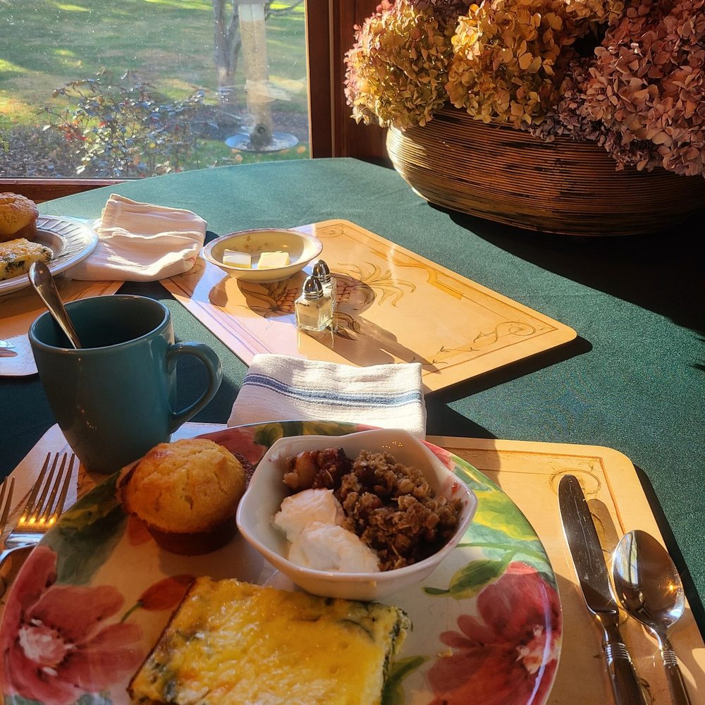 A breakfast table at Journey Inn Bed & Breakfast features a floral plate holding eggs, muffin, and granola, next to a blue mug, utensils, and butter. Sunlight shines in, illuminating dried flowers in a basket by the window.