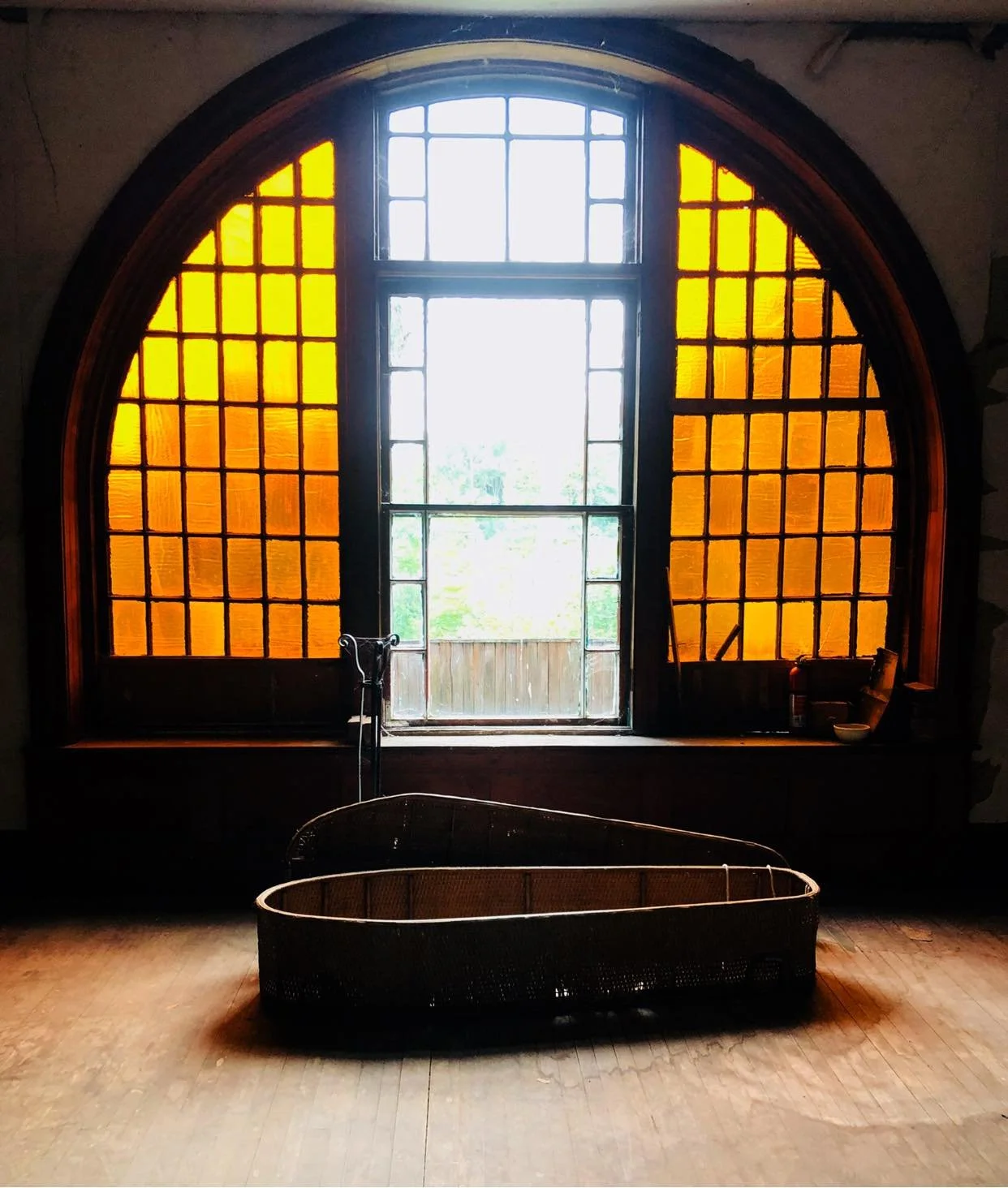A wicker coffin rests on a wooden floor in front of a large arched window with yellow stained glass panels, casting a warm glow into the room—reminiscent of the timeless elegance found in Wilson Castle.