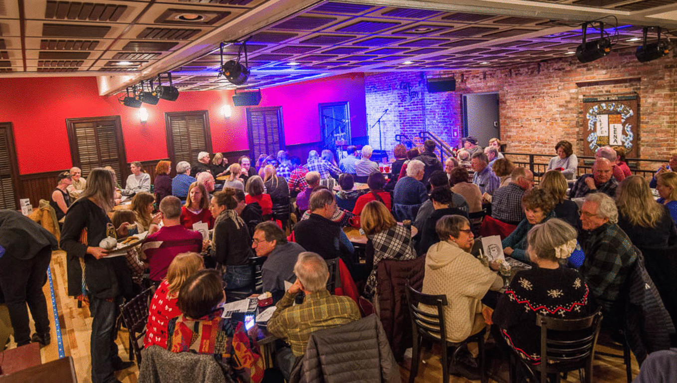 A large group of people sit at tables in a warmly lit venue with red walls and exposed brick, enjoying a social gathering or event that evokes the inviting ambiance of the Schenectady Rose Garden.