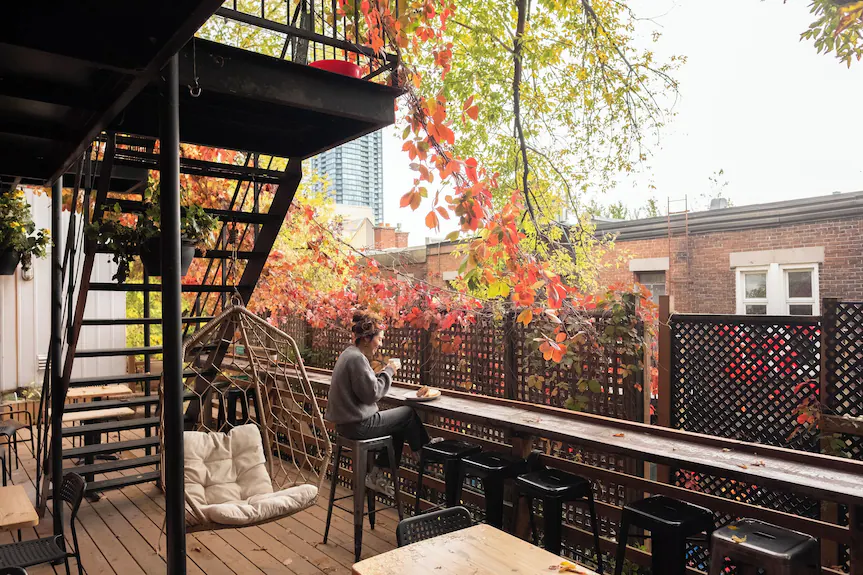 A person sits on a high stool at a wooden patio bar at Auberge Saintlo Montréal, surrounded by autumn leaves and plants, with a metal staircase and nearby buildings visible in the background.