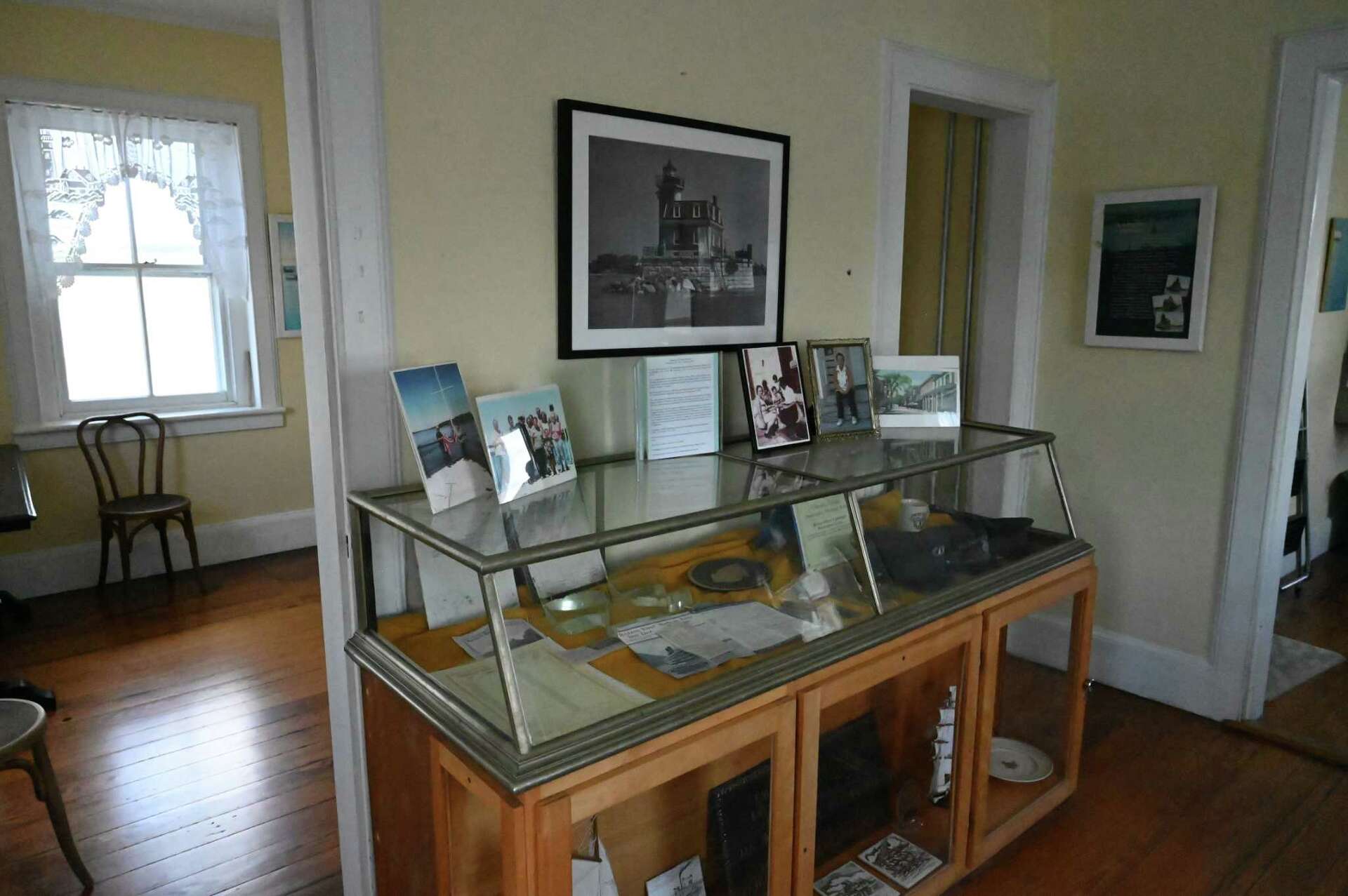 A glass display case with photos, documents, and objects inside a room with wooden floors and light-yellow walls. A framed Hudson-Athens Lighthouse photo hangs above the case, and a window with sheer curtains is on the left.