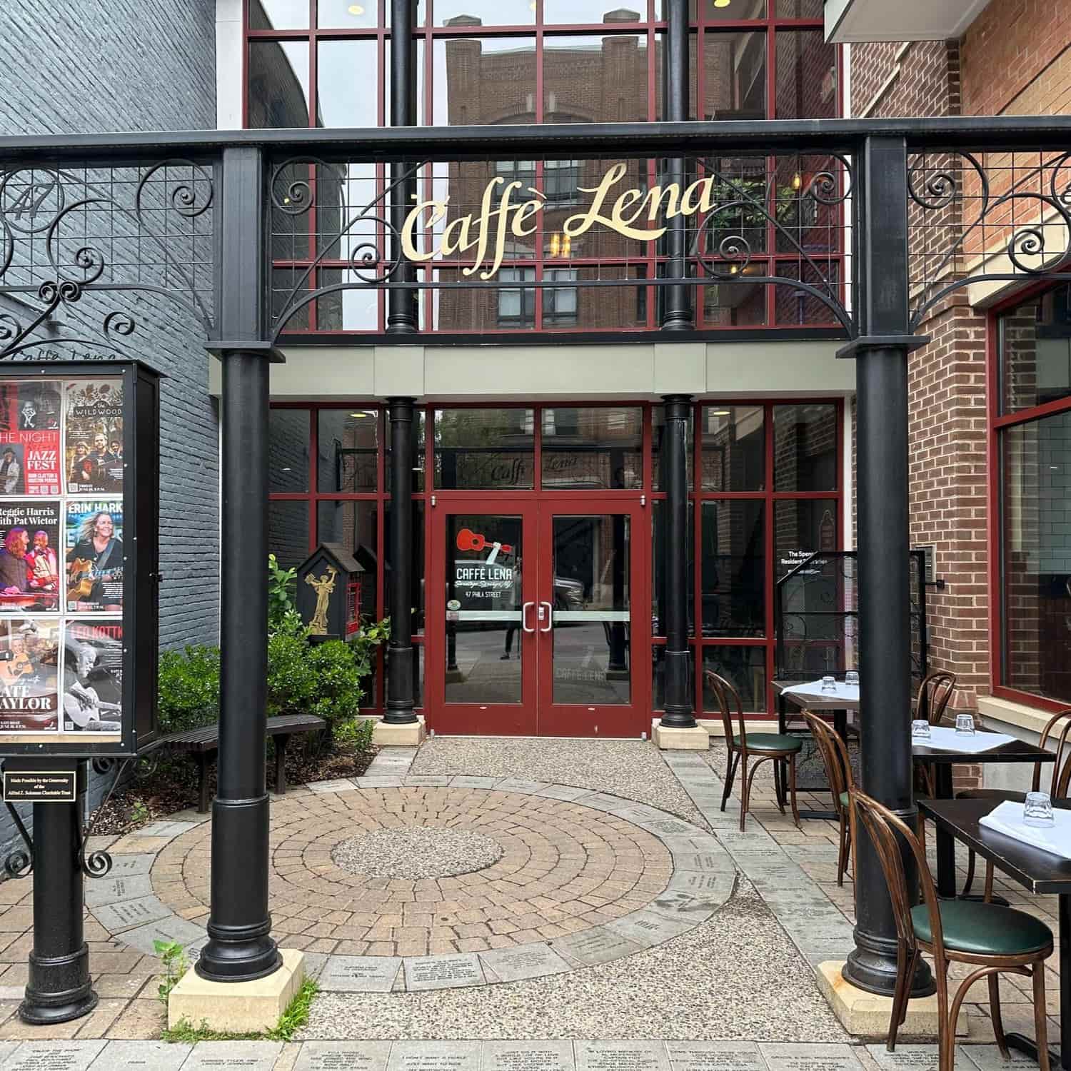 Entrance to Caffe Lena with red double doors, outdoor tables and chairs, black metal archway, and event posters displayed on the left. The paved courtyard and plants evoke a charming atmosphere reminiscent of the Schenectady Rose Garden.