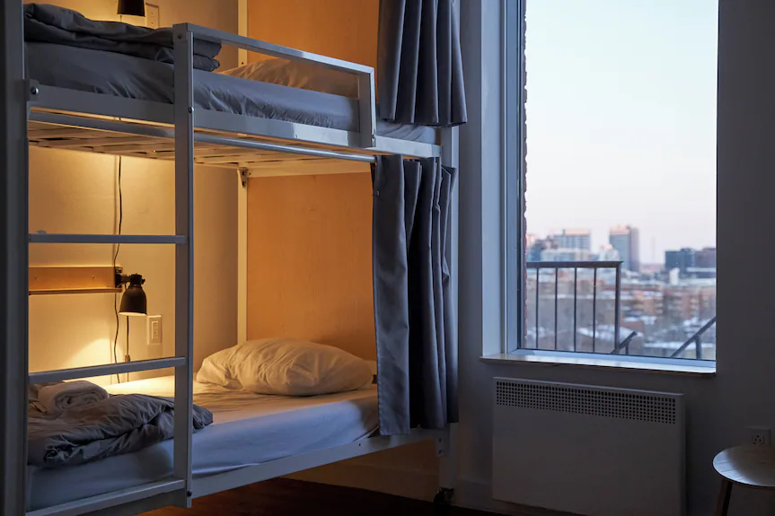 A cozy room at Auberge Saintlo Montréal features bunk beds, one lamp lit, and a large window showcasing a cityscape at dusk. Gray curtains frame both the window and beds, while bedding is crisp white with a pillow and blanket on each mattress.