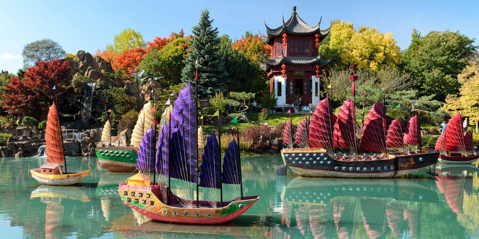 Colorful traditional boats with patterned sails float on a turquoise pond at the Jardin Botanique De Montréal, surrounded by autumn trees and greenery, with a red Chinese pavilion in the background under a clear blue sky.